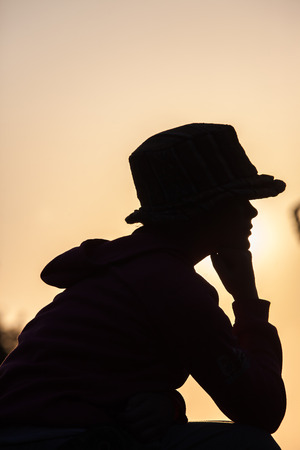 Young teen girl sitting silhouetted body outline wearing hat against sunset dusk sky の写真素材