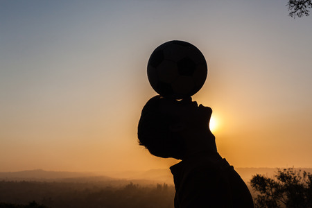 Teenager boy balances football on head silhouetted by sunset dusk skyの写真素材