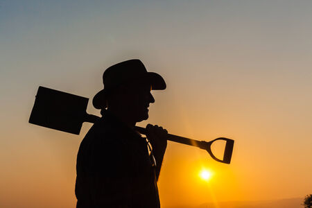 Man holding spade shovel with body outline silhouetted contrasts against sunset dusk colors at days endの写真素材