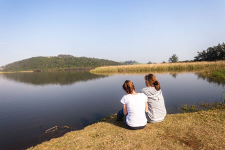 Teen girls relax sitting by beach sea river lagoon waters on a calm blue day in the countrysideの写真素材