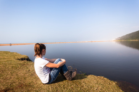 Teen girl relax sitting by beach sea river lagoon waters on a calm blue day in the countrysideの写真素材
