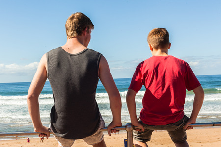 Teen boys family sit on beach railing overlooking blue sea watersの写真素材
