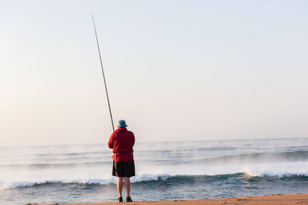 Fisherman surf fishing on beach early morning with river mist out on ocean sea wave waters の写真素材