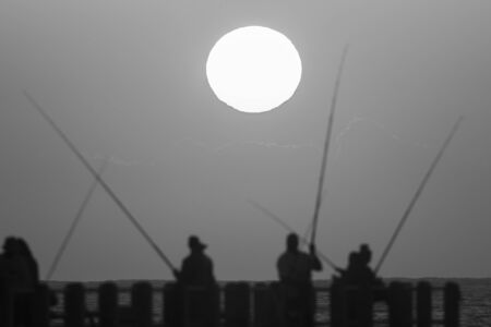 Sunrise fishermen on beach pier silhouetted in black and white vintage.の写真素材