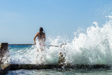 Girl on beach tidal swimming  pool chain wall to facing crashing ocean waves.