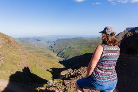 Woman adventurer looks over mountains deep valley dirt road pass landscape summer afternoon.の写真素材
