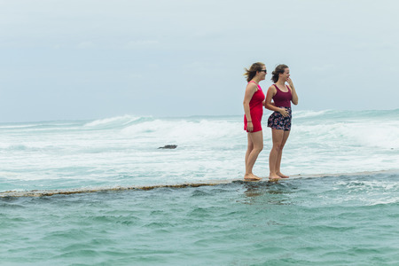 Teenager girls  beach holidays walking talking time on tidal pool wall with overcast ocean waves on horizon.の写真素材