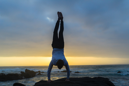 Young man dressed white shirt black longs bare feet gymnastic balance handstand on beach rocks at...