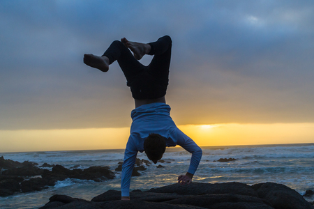 Young man dressed white shirt black longs bare feet gymnastic balance one hand handstand silhouetted...
