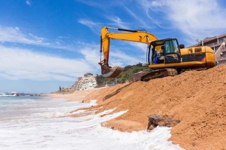 Construction excavators machines on beach water line coastline building repairs with sandbags buffer on sand dunes to protect road from ocean storms.のeditorial素材