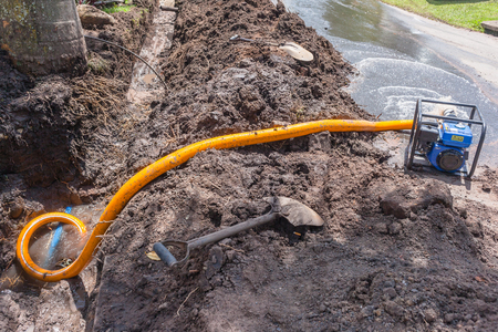 Plumbing pumping water from earthwork trench digging repairs of broken main  pipe in residential road area with mobile portable motor pump draining damage.の写真素材