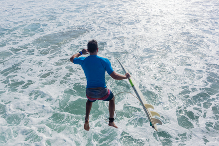 Surfer with surfboard jumps off beach pier jetty into ocean water for quick entry to go surfing ...