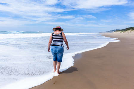 Caucasian Mature woman holidays walking beach ocean shoreline with wave foam wash onto her feet a rear unidentified photo along coastal countryside landscapeの写真素材