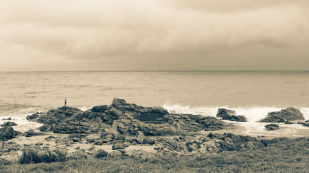 Lifestyle distant fisherman on beach coastline rocks fishing with distant rain storm coming over the ocean seas a panoramic sepia tone photo landscape.の写真素材
