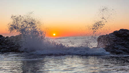 Dawn beach rocky tidal pool with wave crashing water spray against ocean horizon sunrise.