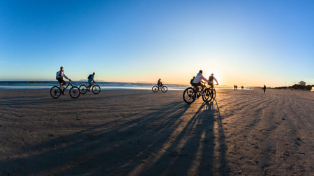 Beach ocean coastline with riders bicyles cycling silhoettes as sun dipping  behind mountains  a scenic lifestyle.の写真素材