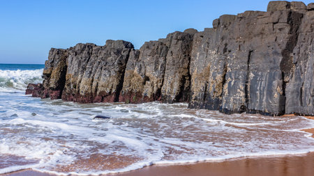 Beach black rock wall headland into sand on waters edge towards blue ocean waves and horizon blue sky landscape.の写真素材