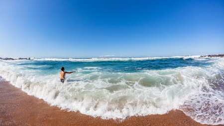 Young caucasian boy on beach sand enters ocean into crashing shorebreak waves for a swim , a rear behind photograph on a summer blue day.の写真素材