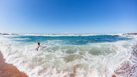 Young caucasian boy on beach sand enters ocean into crashing shorebreak waves for a swim , a rear behind photograph on a summer blue day.の写真素材