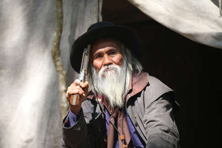 Old rough western cowboy with gray beard and brown hat holding rifle.