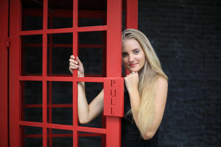 Portrait of Beautiful blonde hair girl on black dress standing in red phone booth against black wall as portrait fashion pose outdoor.の写真素材