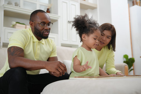 Shot of joyful parents with their little daughter sitting on floor inside living room in sunny day.....