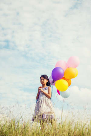 Cute little girl holding colorful balloons in the meadow against blue sky and clouds,spreading hands.の写真素材