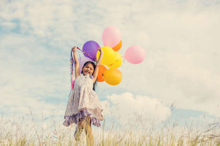 Cute little girl holding colorful balloons in the meadow against blue sky and clouds,spreading hands.の写真素材