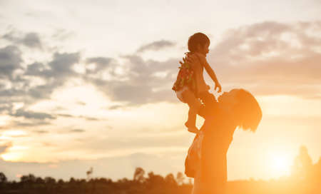 a silhouette of a happy young girl child the arms of his loving mother for a hug, in front of the sunset in the sky on a summer day.の写真素材