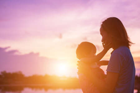 a silhouette of a happy young girl child the arms of his loving mother for a hug, in front of the sunset in the sky on a summer day.の写真素材