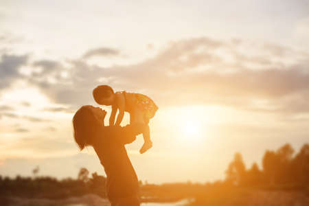 a silhouette of a happy young girl child the arms of his loving mother for a hug, in front of the sunset in the sky on a summer day.の写真素材