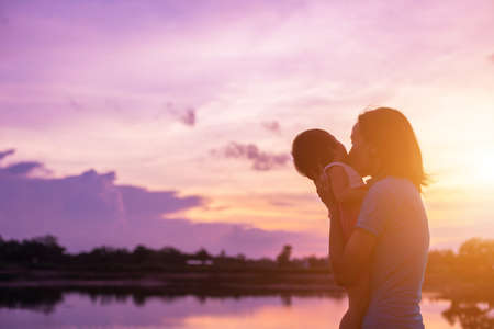 a silhouette of a happy young girl child the arms of his loving mother for a hug, in front of the sunset in the sky on a summer day.の写真素材