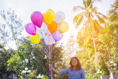 young asian woman running and jumping on green grassland with colored balloonsの写真素材