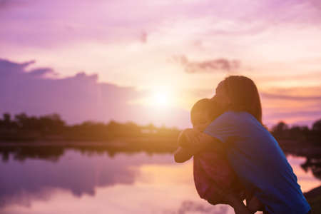 a silhouette of a happy young girl child the arms of his loving mother for a hug, in front of the sunset in the sky on a summer day.の写真素材