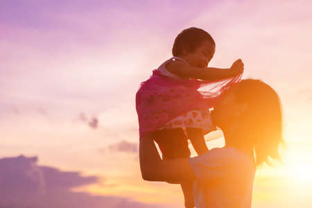 a silhouette of a happy young girl child the arms of his loving mother for a hug, in front of the sunset in the sky on a summer day.の写真素材