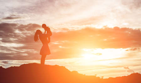 a silhouette of a happy young girl child the arms of his loving mother for a hug, in front of the sunset in the sky on a summer day.の写真素材