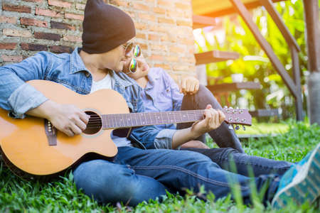 Hipster man playing guitar for his girlfriend outdoor against brick wall, enjoying together.の写真素材