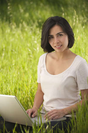 beautiful, young Hispanic woman working on a laptop computer in a grassy meadowの写真素材