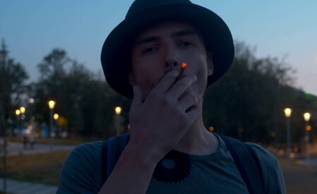 Portrait of a young man smoking a cigarette on the night city park backgroundの写真素材