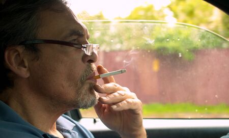 Close up portrait of handsome senior man driving a car and smoking. Shooting inside the carの写真素材