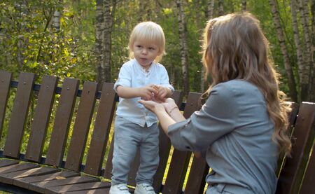 Toddler standing on the bench and eating cookies. Mother sitting nearby. Sunny day in the park