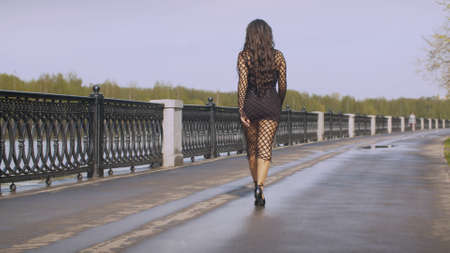 Beautiful fashionably dressed woman walking along the promenade in a city. Nice early spring day, fresh leaves on the trees. Real people. Follow shotの写真素材