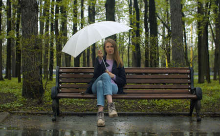 Portrait of a beautiful pensive woman with umbrella sitting on the bench in the park in spring. Rainy weather, real peopleの写真素材