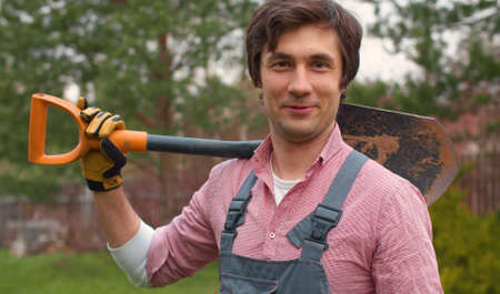 Portrait of a gardener with a shovel on his shoulder. Smiling young caucasian man in overalls in the...
