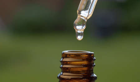 Macro shot essential oil dripping from a pipette on blurry natural background. Extreme close up hand...