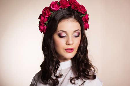 Fashion portrait of cute teenager girl with nice makeup is wearing white blouse and red roses wreath on her headの写真素材