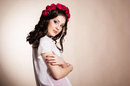 Fashion portrait of happy pretty brunette teenager girl is wearing white blouse and red roses wreath.の写真素材
