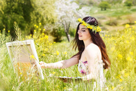 Young female artist is wearing wreath of yellow flowers is create picture of an floral landscape, spring timeの写真素材