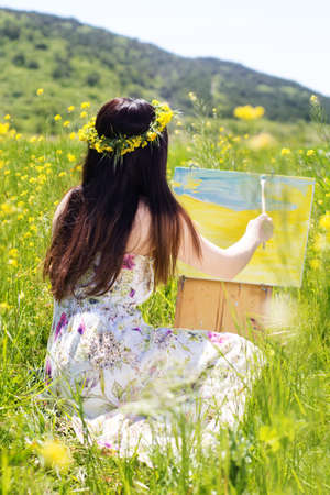 Young female woman is wearing wreath of yellow flowers is painting picture of an flowers and mountains, spring timeの写真素材