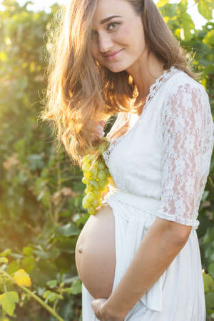 Happy pregnant girl is wearing white lace fashion dress is holding bunch of grapes near her belly in...